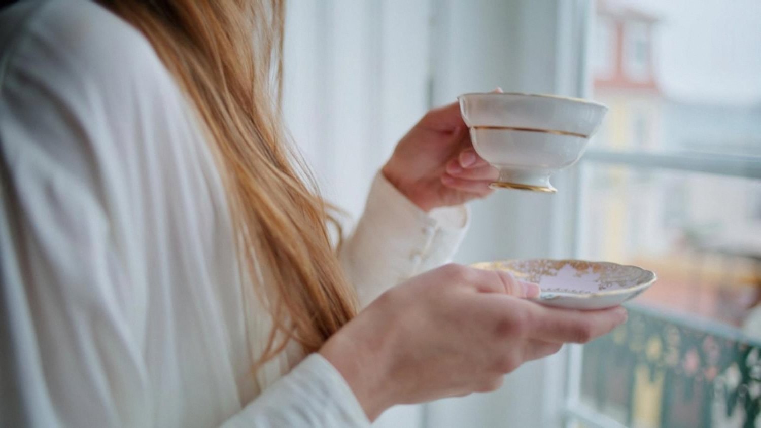 Woman holding a delicate porcelain cup and saucer near a sunlit window at home