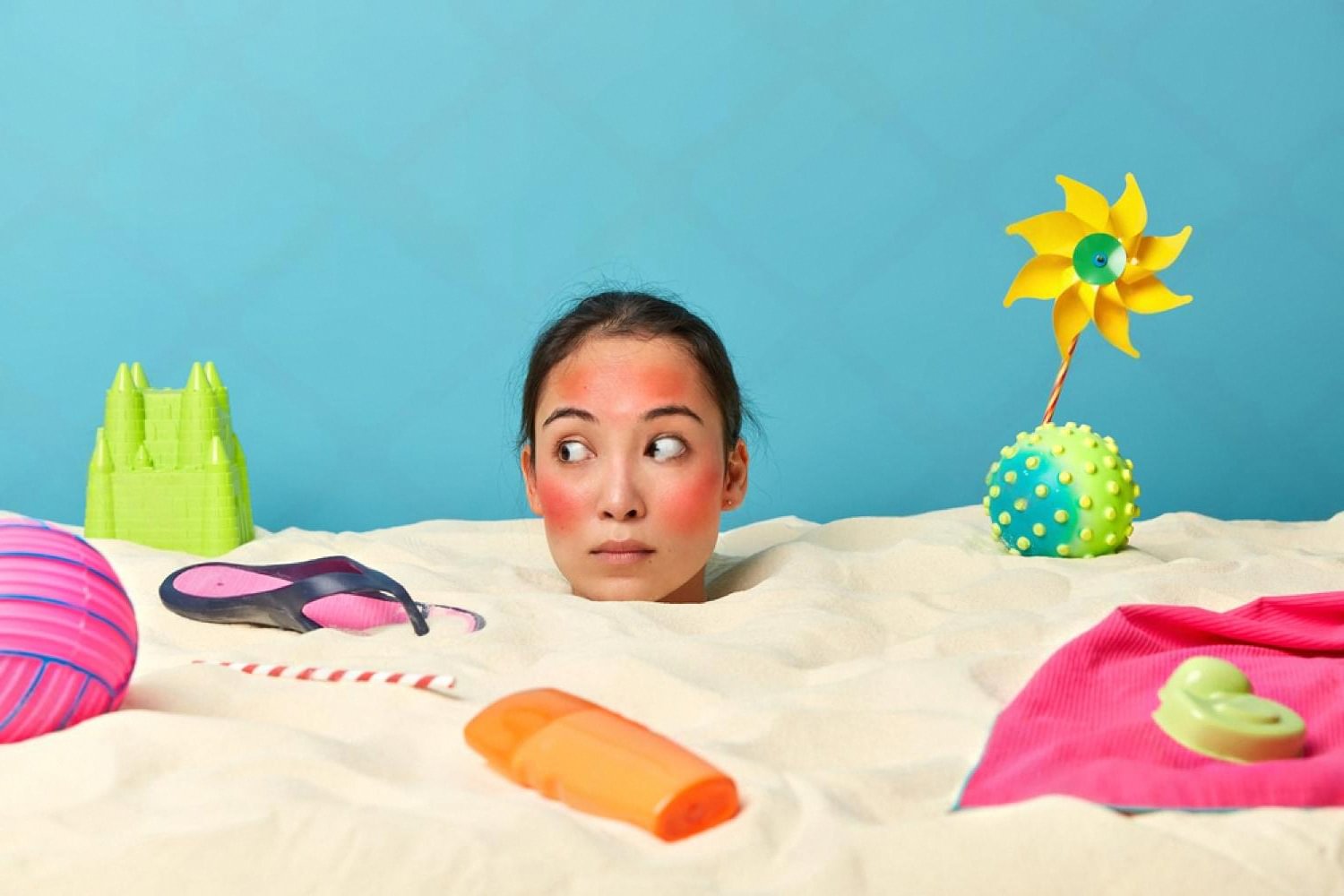 Woman with sunburned red cheeks buried in beach sand surrounded by summer items illustrating UV damage to the skin barrier